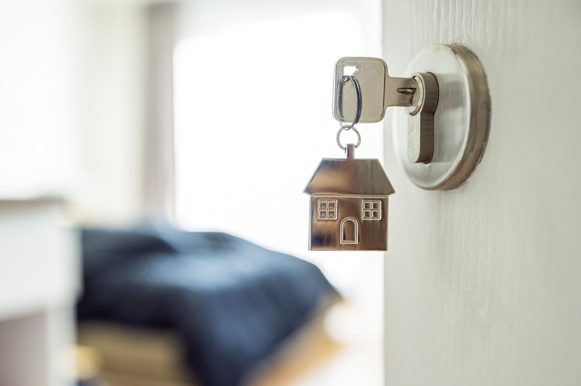 Key in a door with a house-shaped keychain, bedroom in background.