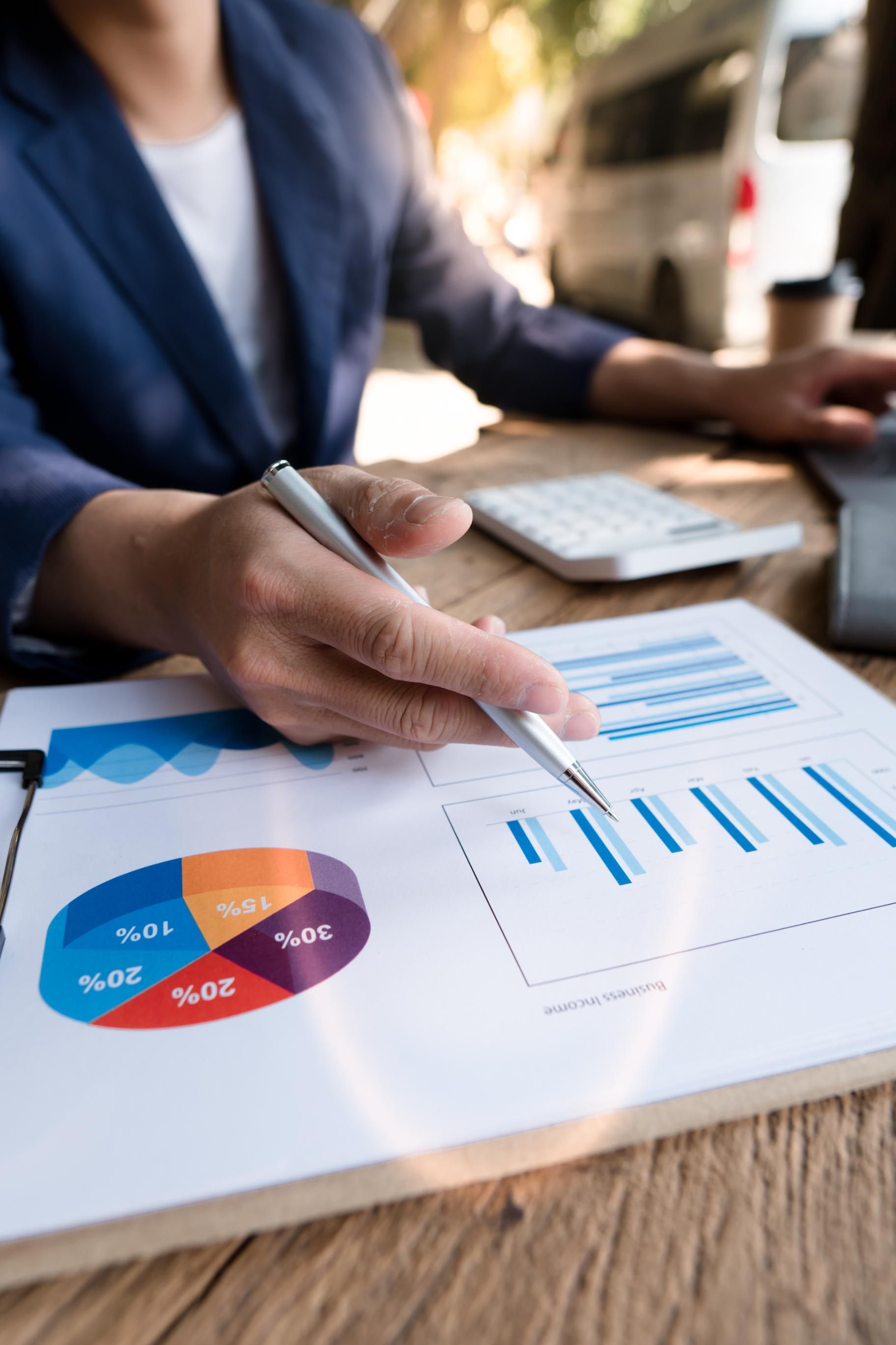 Person in blue blazer reviews charts with pen at a wooden desk, including a pie chart and bar graph.