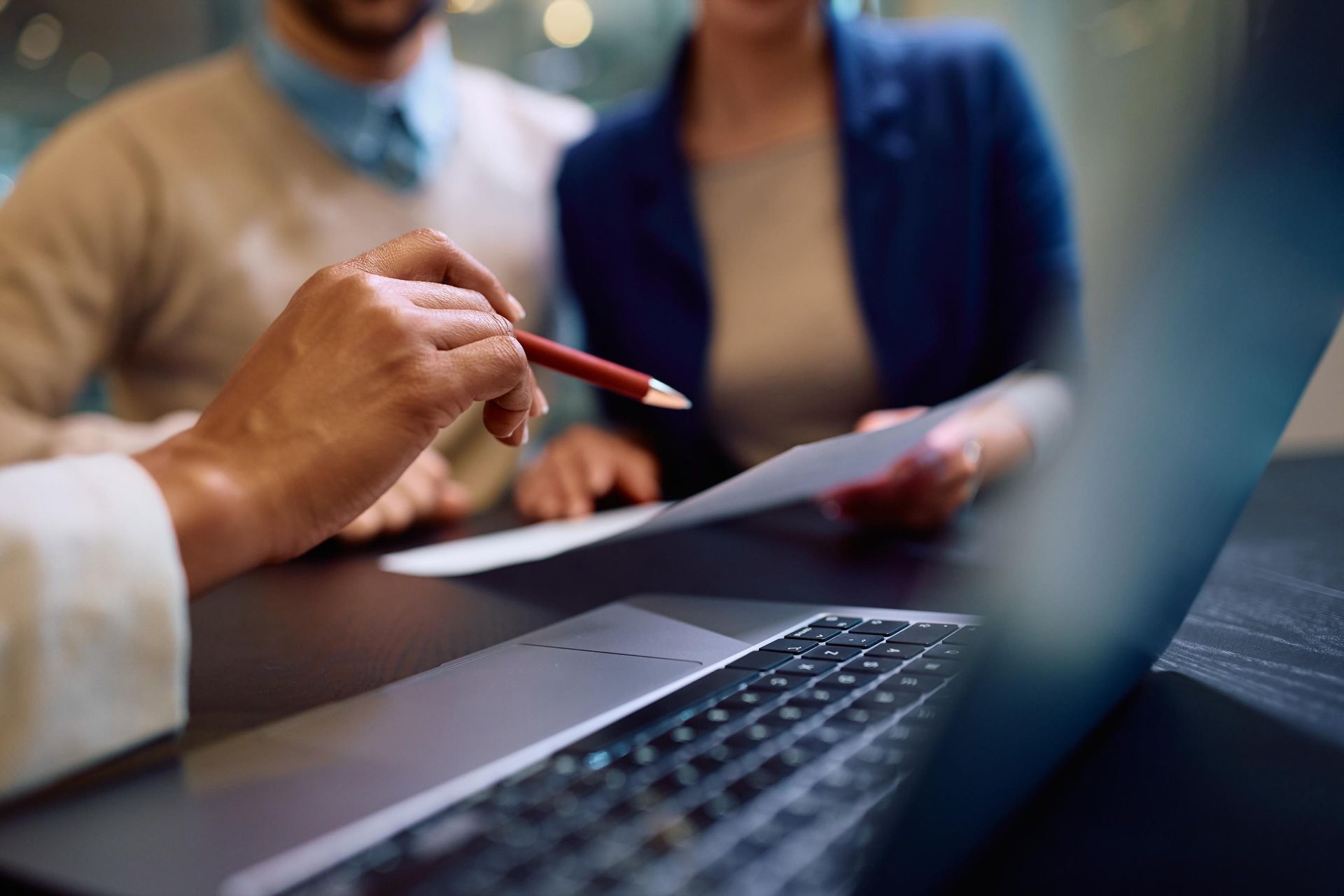 A person pointing a pen at a document; couple looks on. Laptop sits on table.