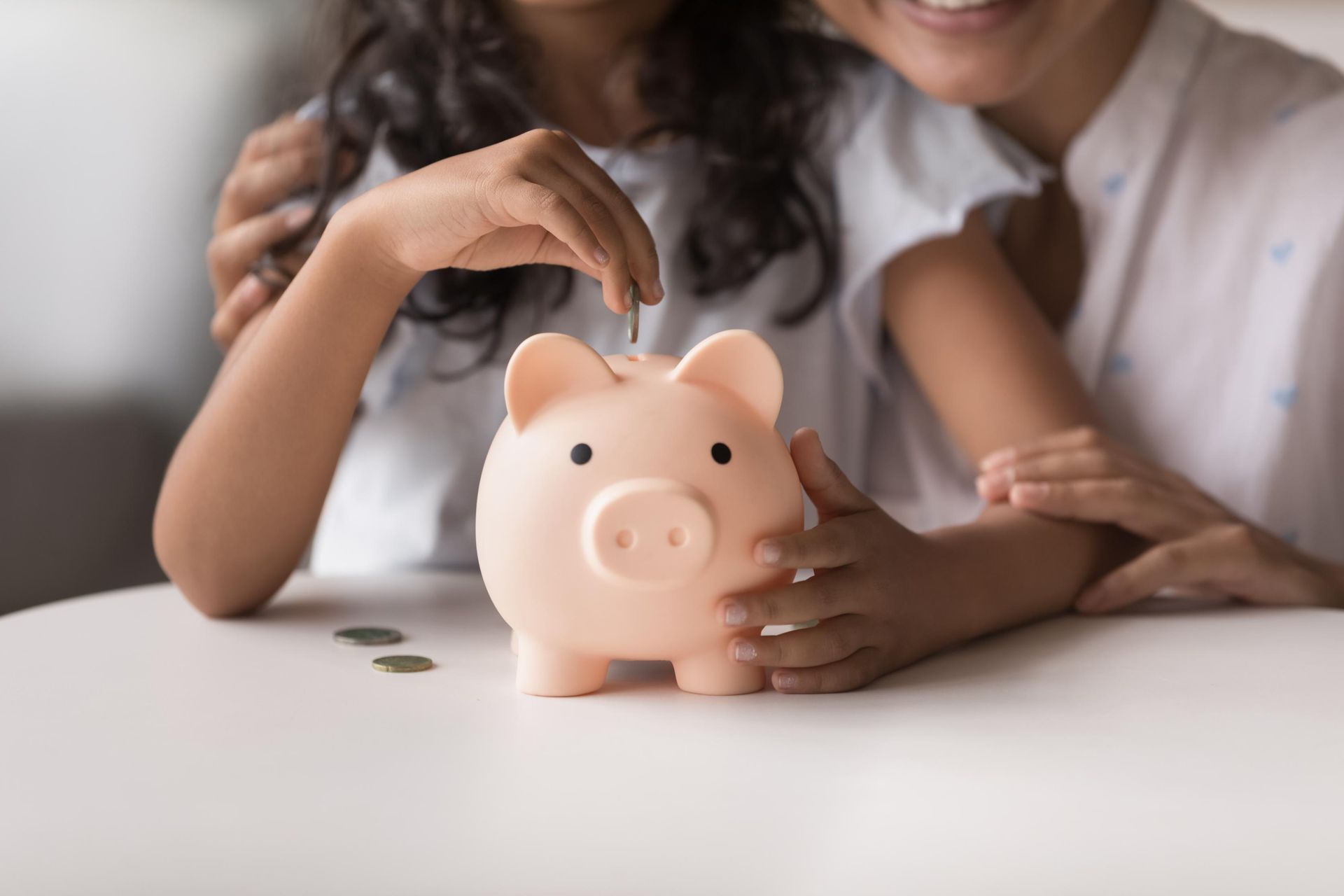 Child putting a coin in a pink piggy bank with parent's hand on her arm, smiling.
