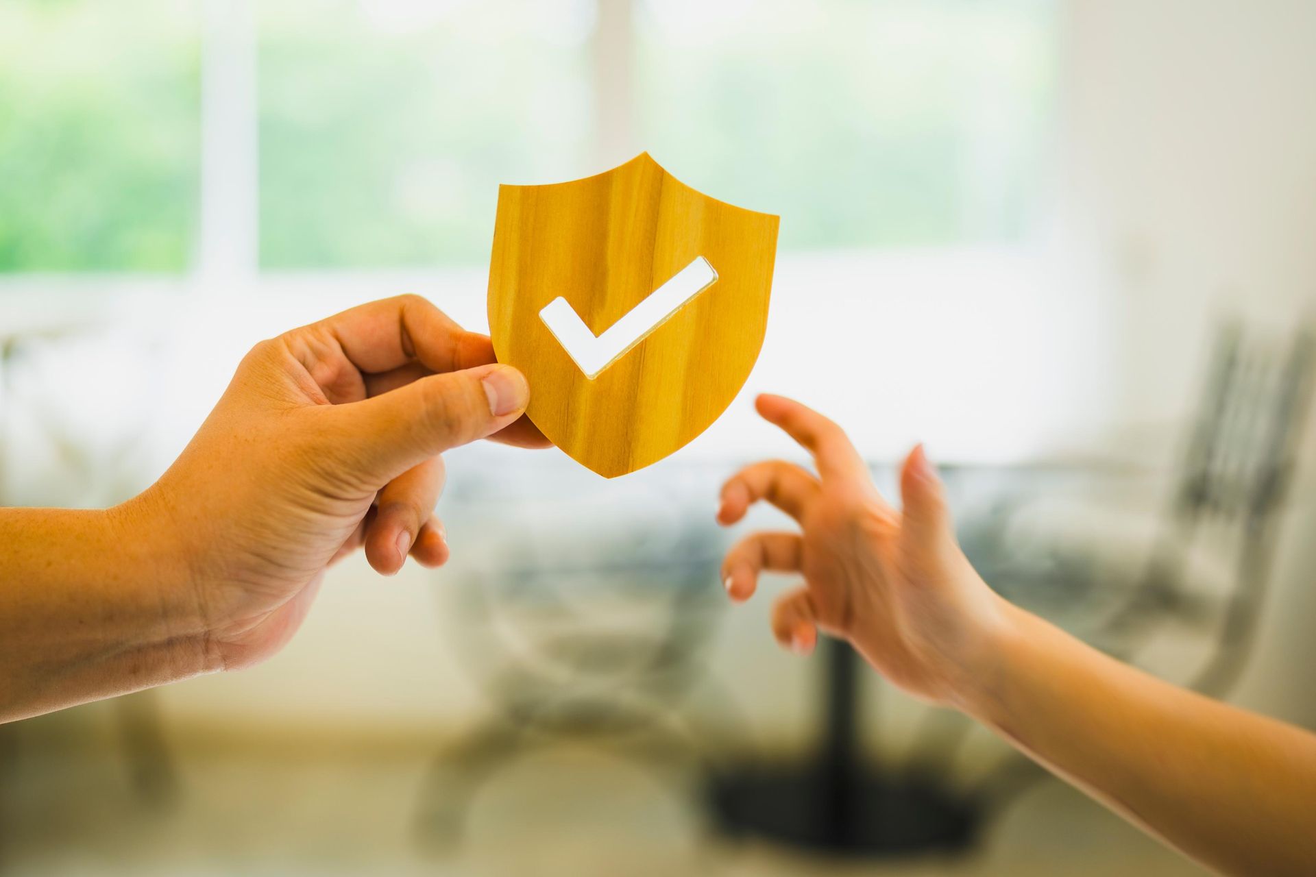 Hands holding up a yellow shield with a white checkmark, with a blurred background of a room.