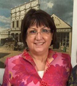 A woman wearing glasses and a pink jacket is smiling in front of a painting.