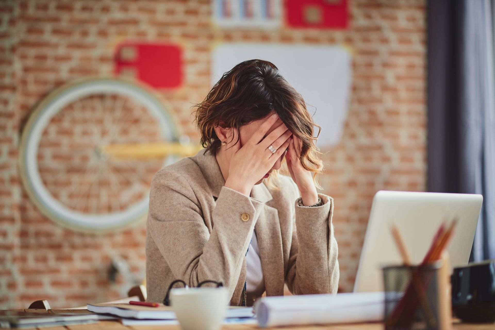 Person covering face at a desk with laptop and papers, appearing stressed in a brick-walled office