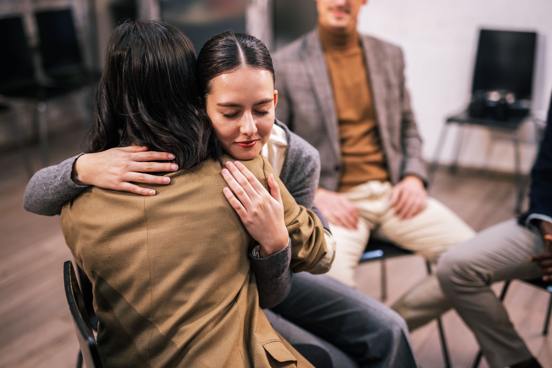 Two people sit closely in a support group, one comforting the other with an embrace.