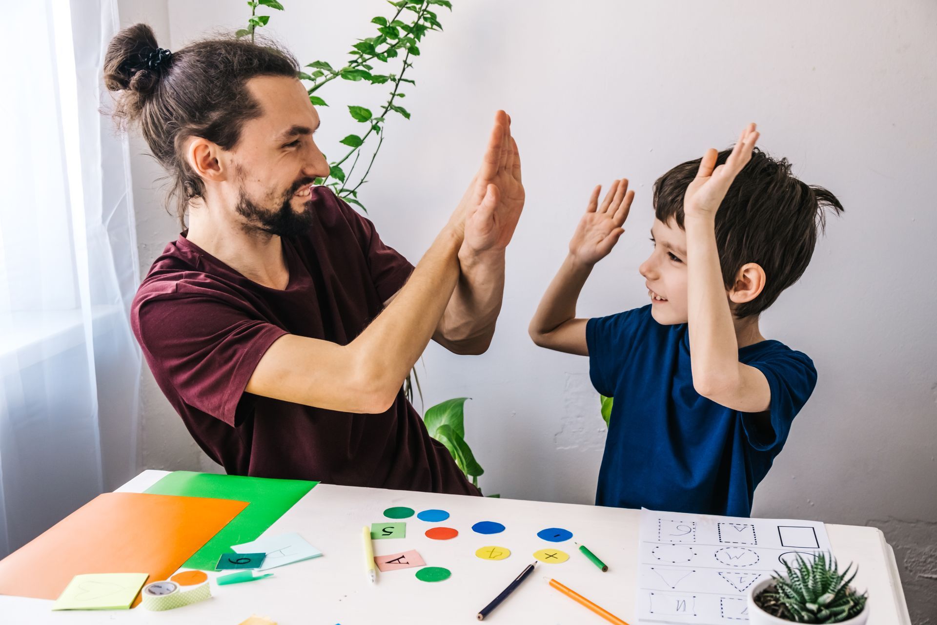 Man and child high-fiving at a table with craft supplies in a bright room