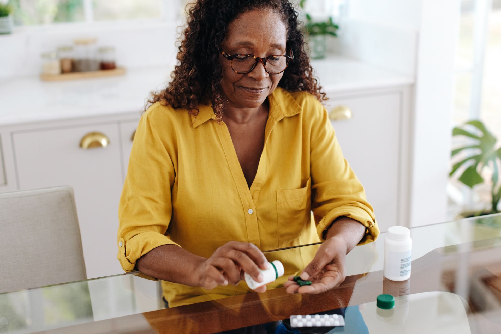 Woman in yellow shirt taking medication at a glass table, with pill bottles nearby