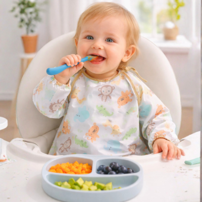 Bebé en una trona comiendo con cuchara y con babero. Plato de comida visible.