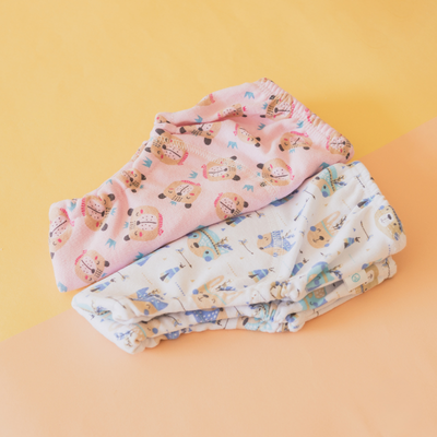 Dos pares de ropa interior de bebé, uno rosa y otro blanco, ambos con estampado de osito, sobre fondo naranja y amarillo.