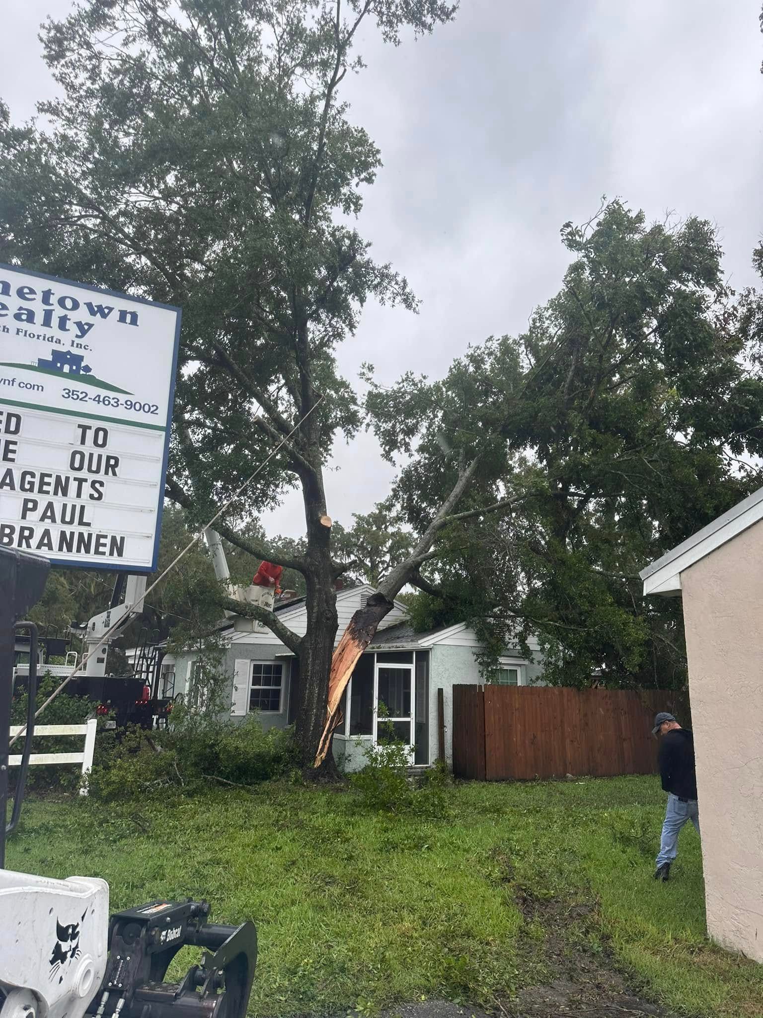 A man is standing in front of a house with a tree fallen on it.