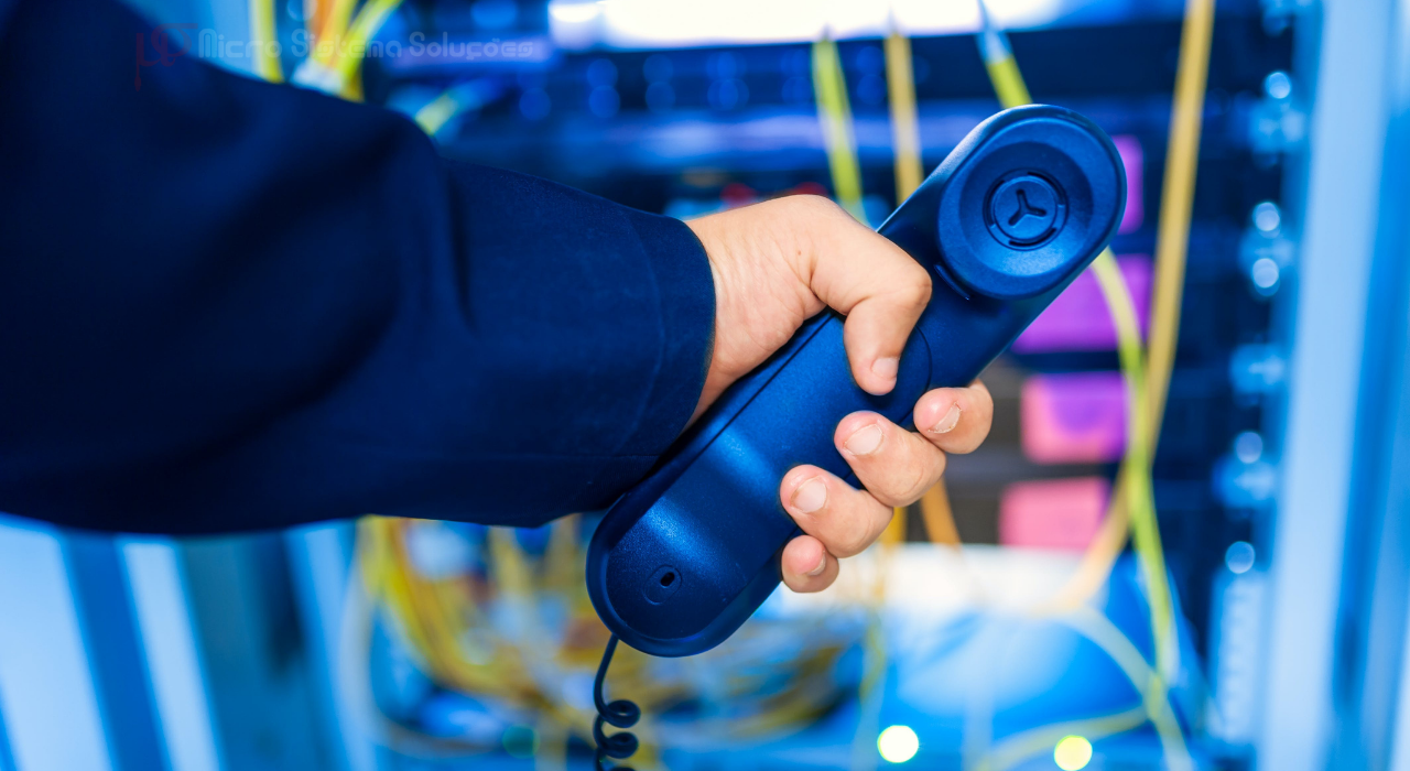 A hand holding a blue phone receiver in front of a server rack with colorful cables.