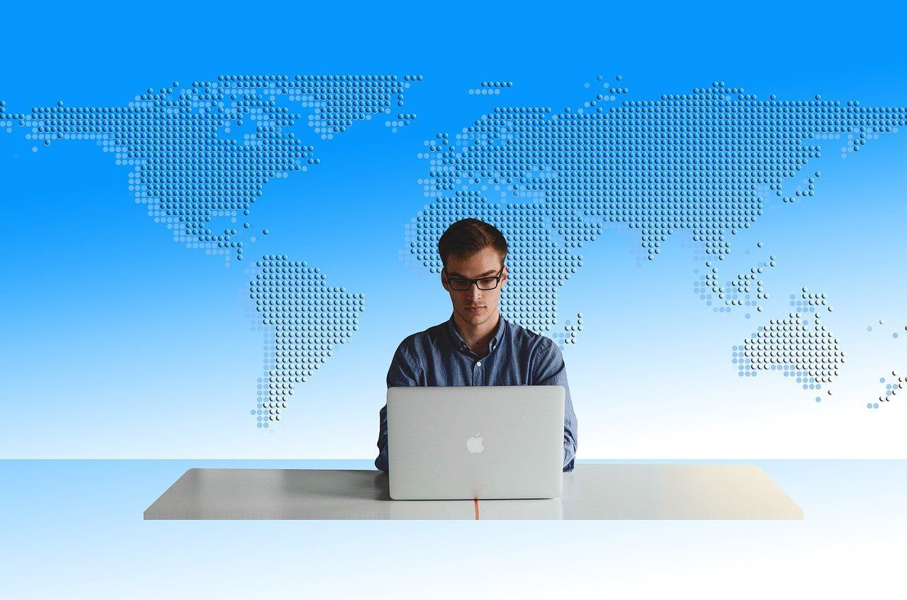 Um homem está sentado em uma mesa usando um laptop em frente a um mapa do mundo.