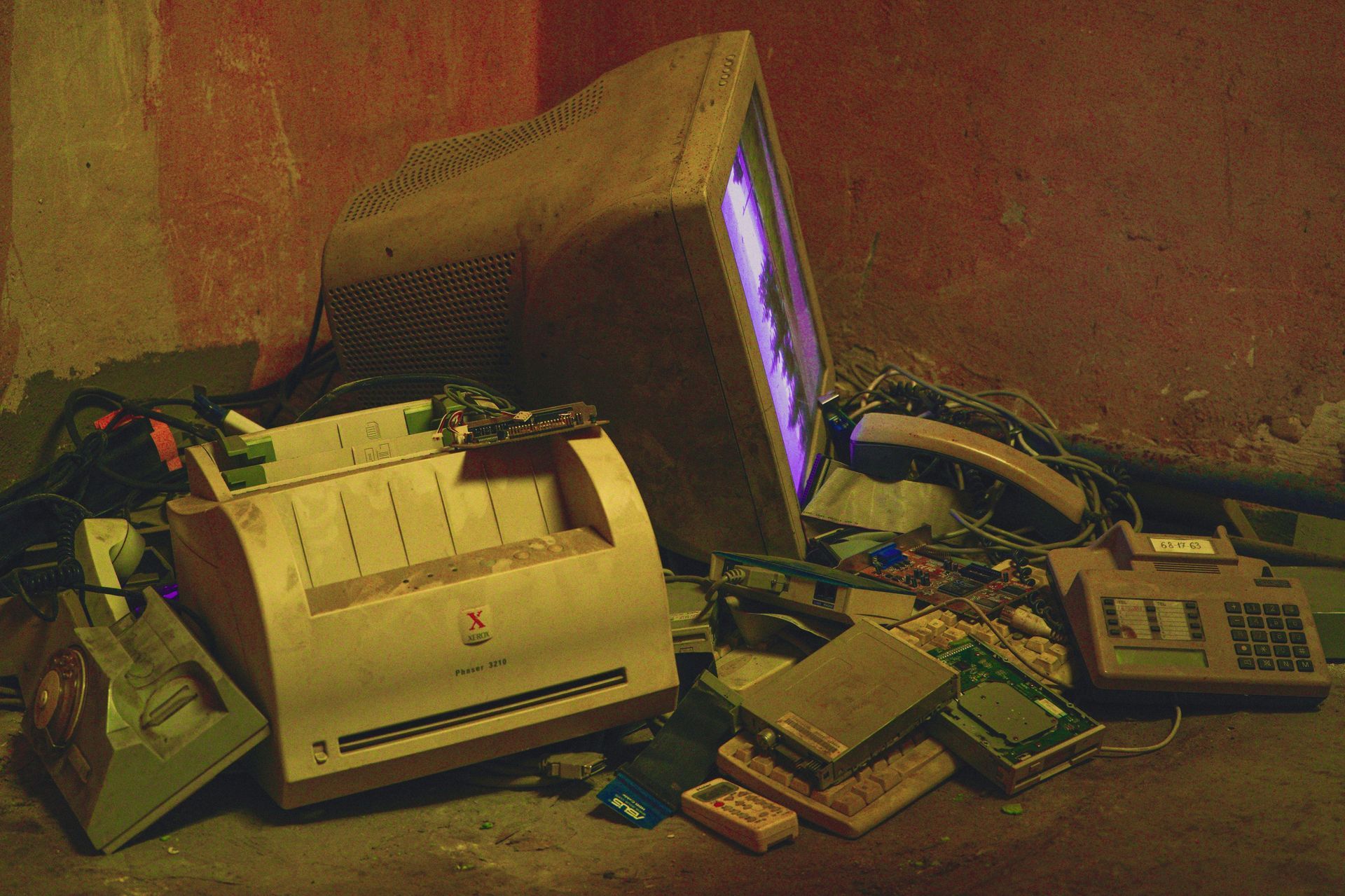 Heap of obsolete electronics: CRT monitor, printer, circuit boards, cables, in a dimly lit space.