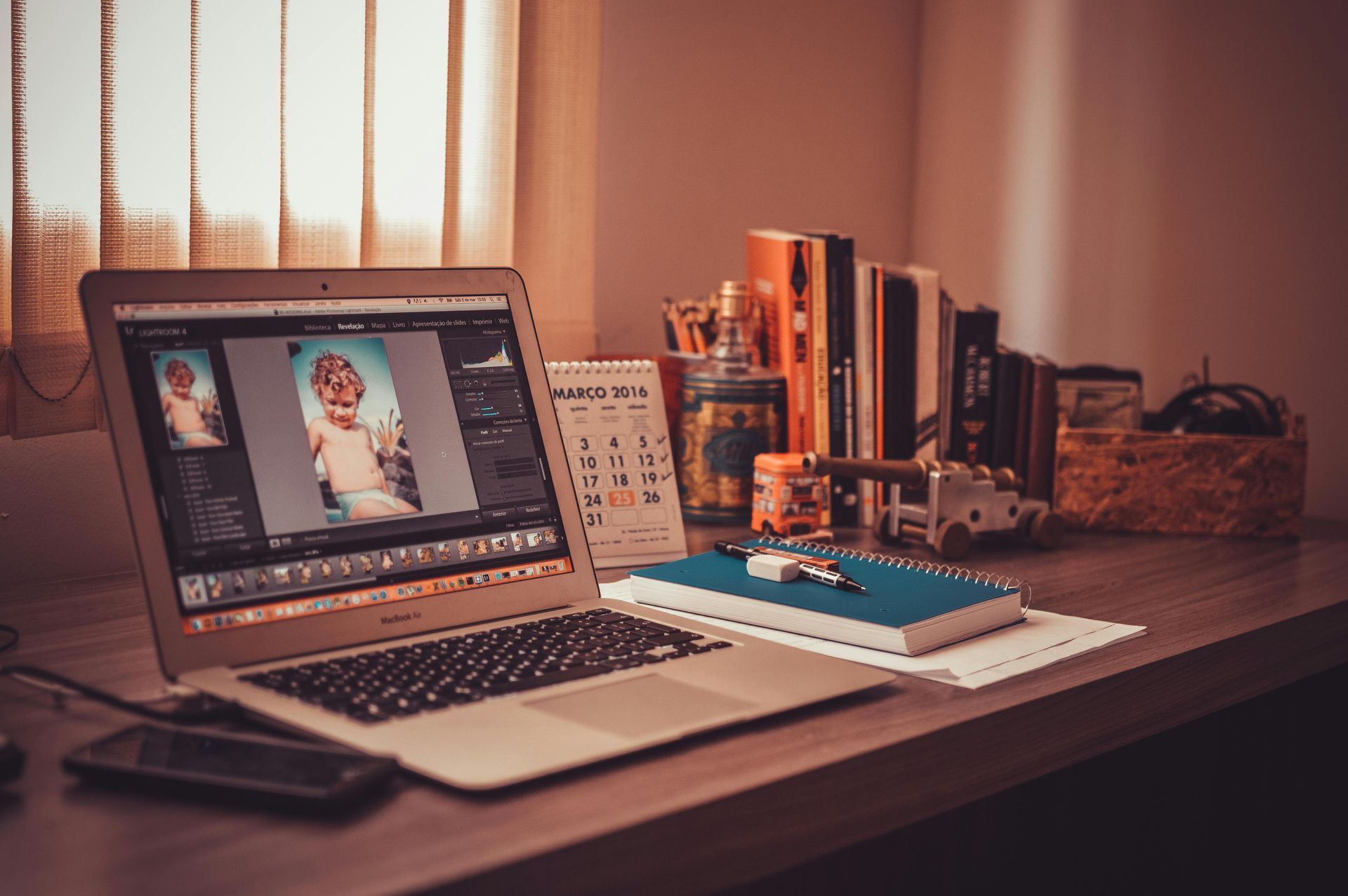 Laptop displaying photo editing software on a desk with books, calendar, and a phone.
