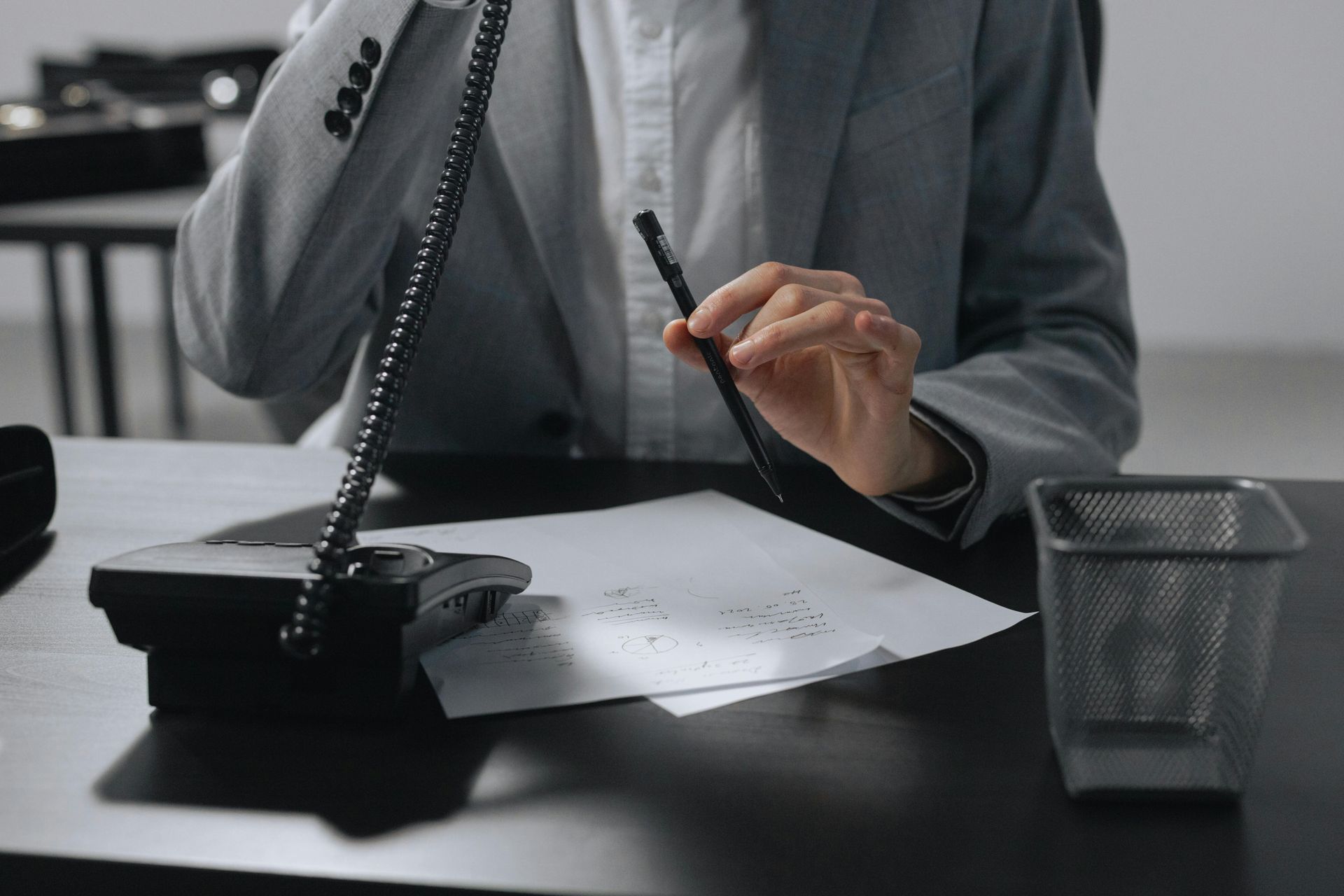 Person in suit on the phone, holding a pen, looking at a paper on a desk.