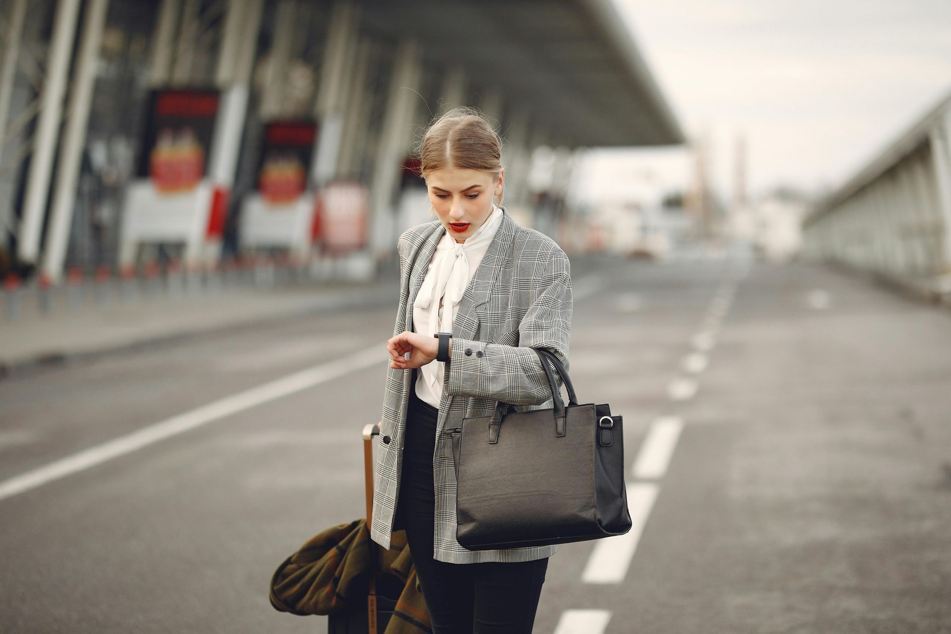 Woman in business attire at airport, looking at watch. Holding bag and suitcase, coat over arm.