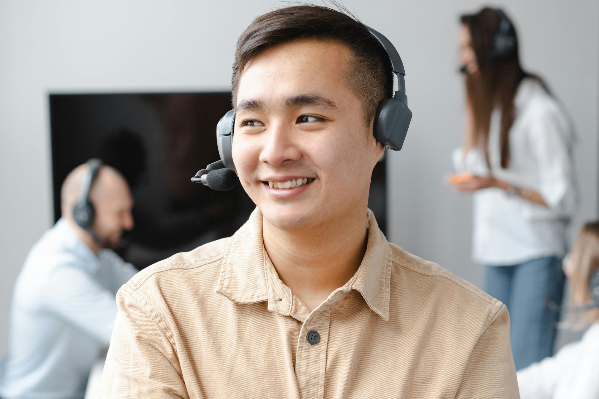 Man with headset smiles, looking to the side; colleagues in background.