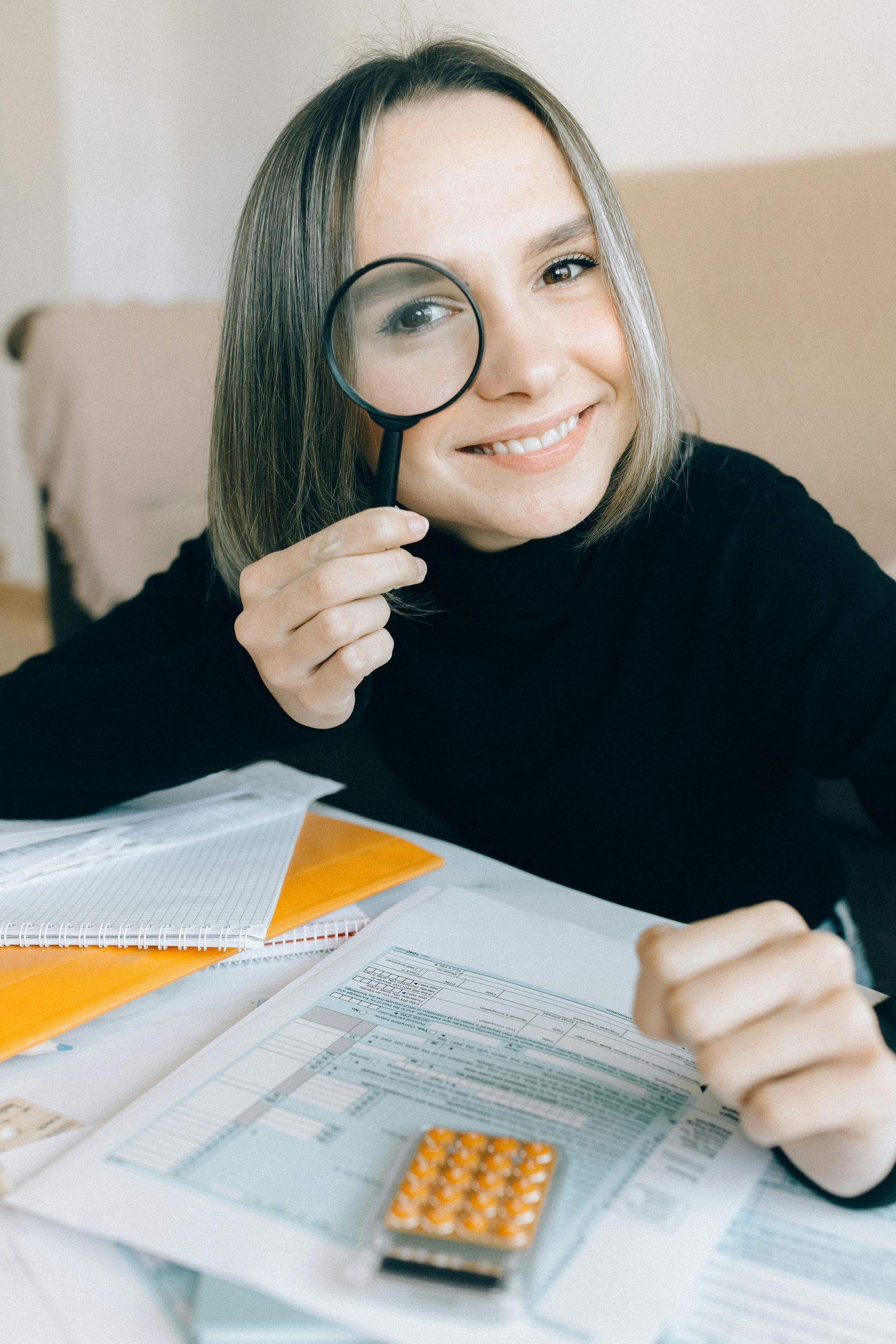 Woman smiling, peering through magnifying glass at documents, calculator nearby.