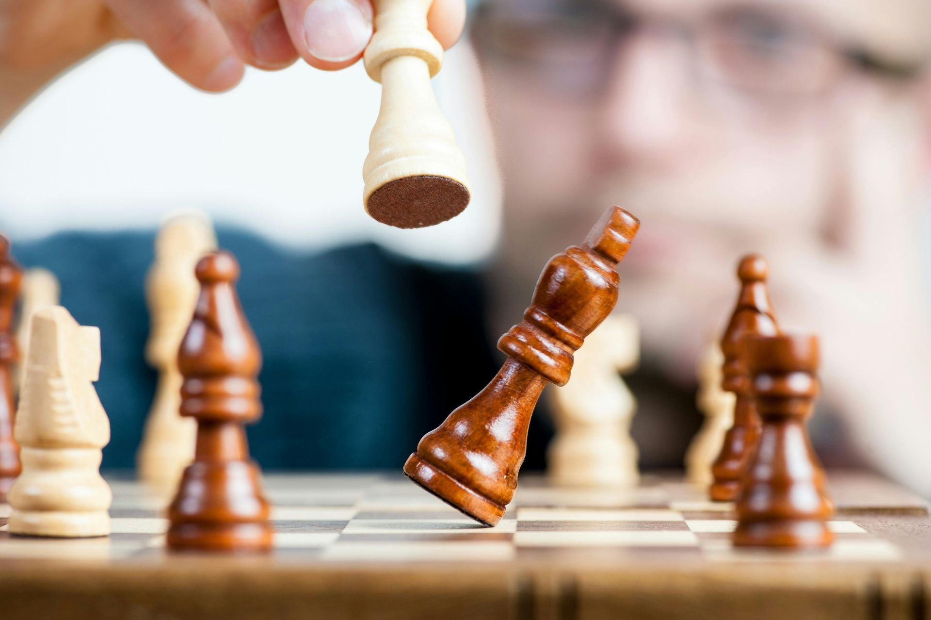 A person moves a white chess piece toward a fallen brown king on a chessboard.