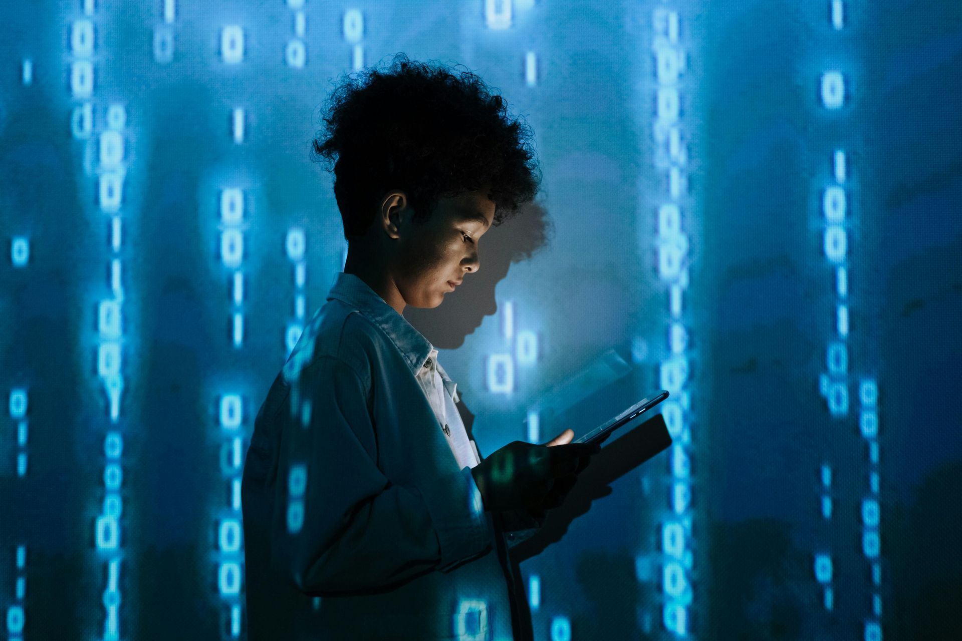 Person with afro looking at a tablet, lit by blue binary code in a dark room.