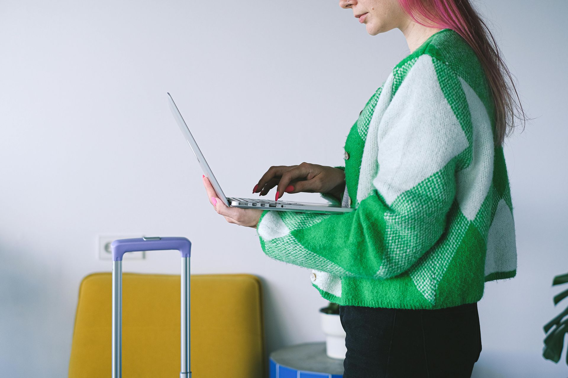 Person typing on a laptop, standing near luggage and a yellow seat; wearing a green and white sweater.