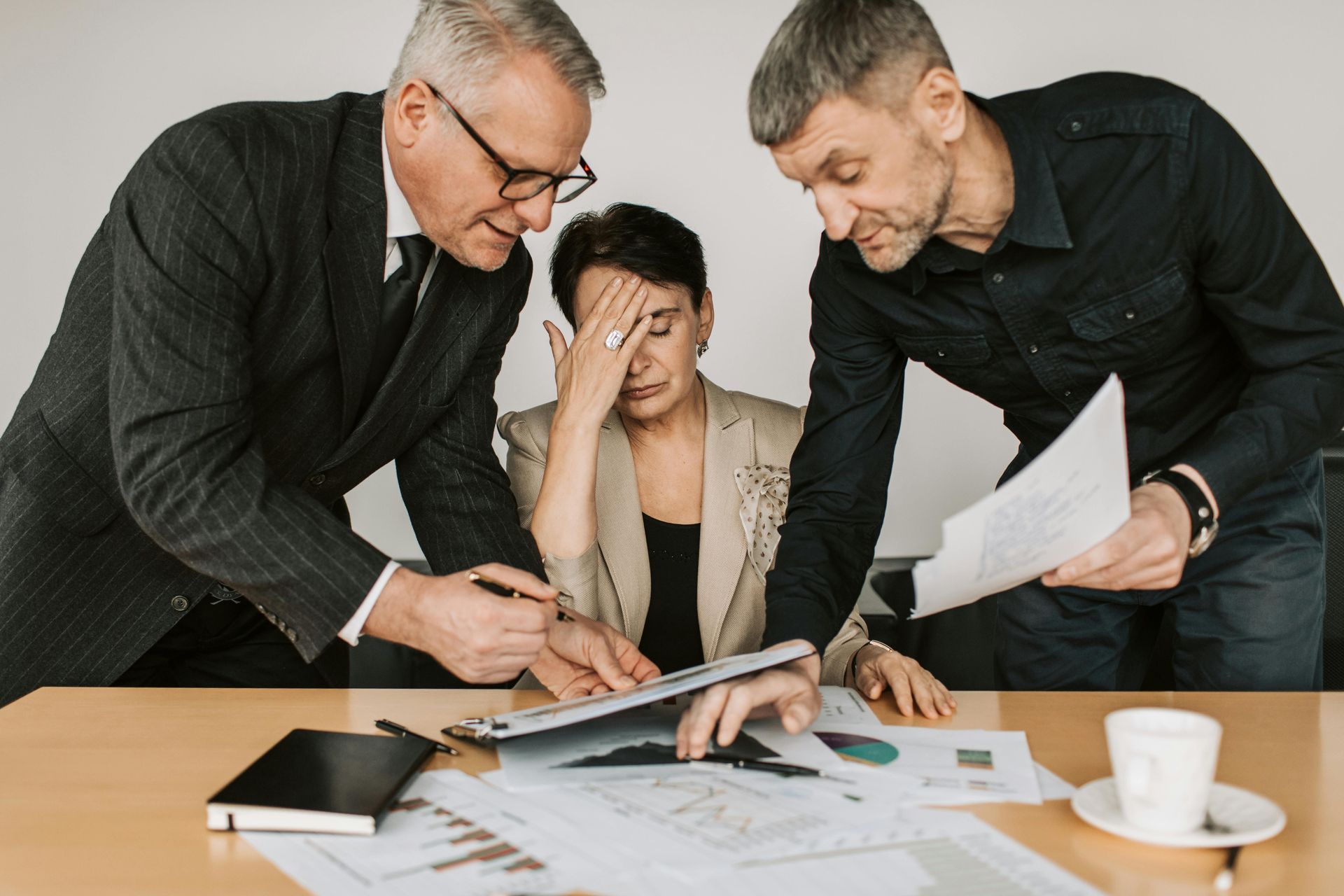 Three people at a desk reviewing documents; a person sits with their hand on their forehead while two others stand nearby.