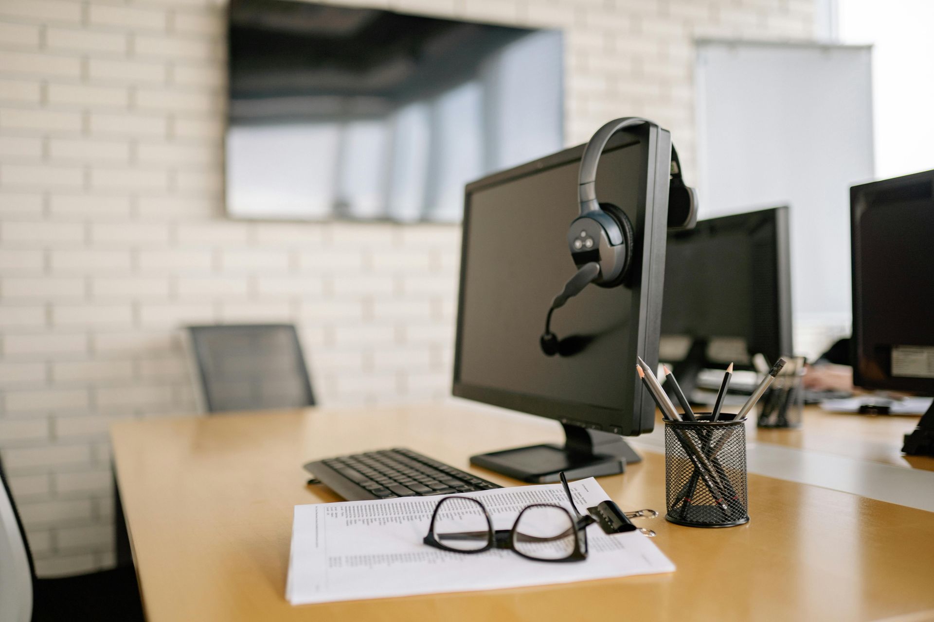 Desk with computer, headset, keyboard, glasses, and documents in a modern office setting.