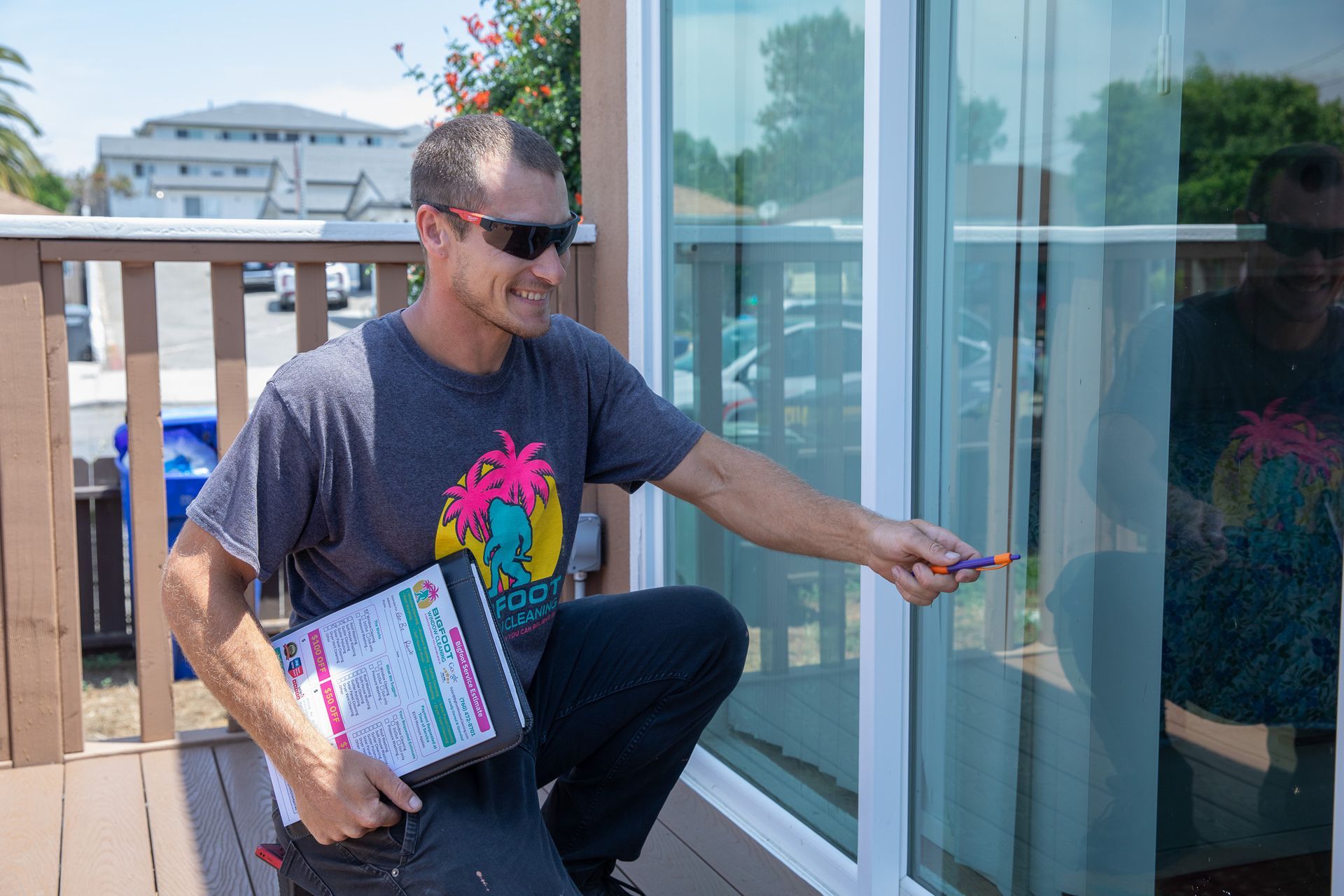 A man is kneeling on a deck holding a clipboard and pointing at a window.