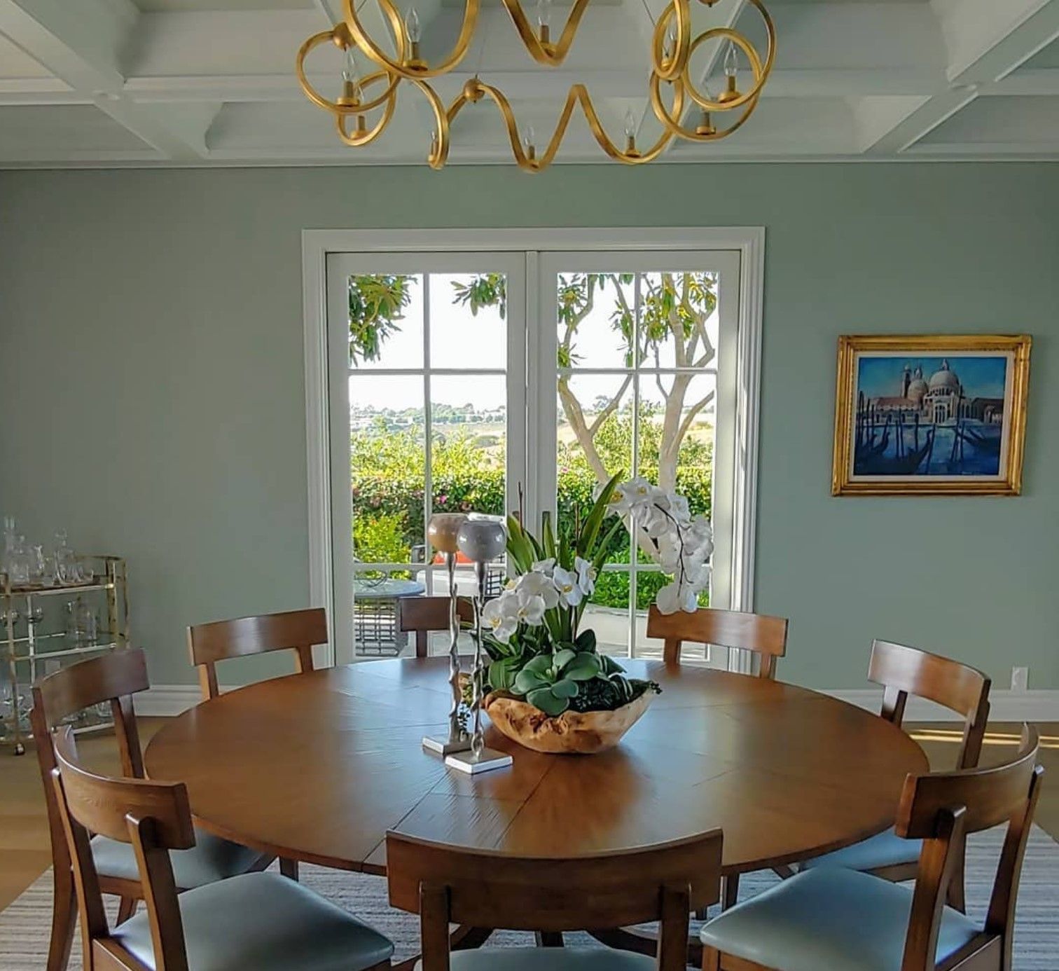 Dining room with round wooden table, chairs, and a chandelier. A painting hangs on the wall;