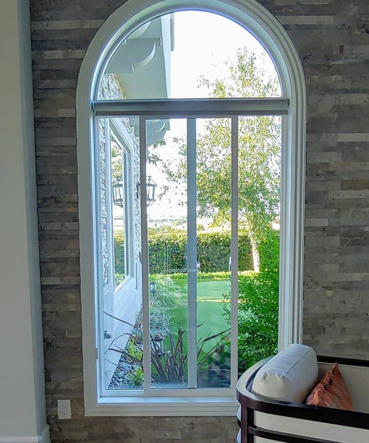 Arched white window with view of a garden, with a stone wall on the side and a curved chair at the right.