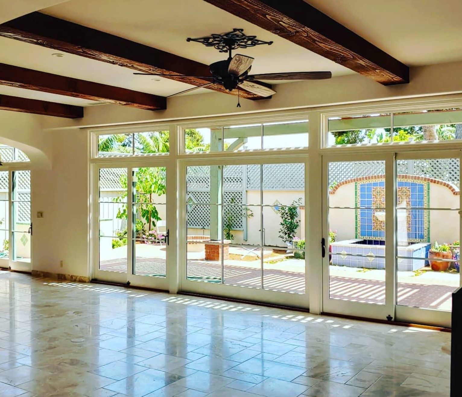 Sunlit room with large white-framed glass doors opening to a courtyard. Exposed wood beams on ceiling, marble floor.