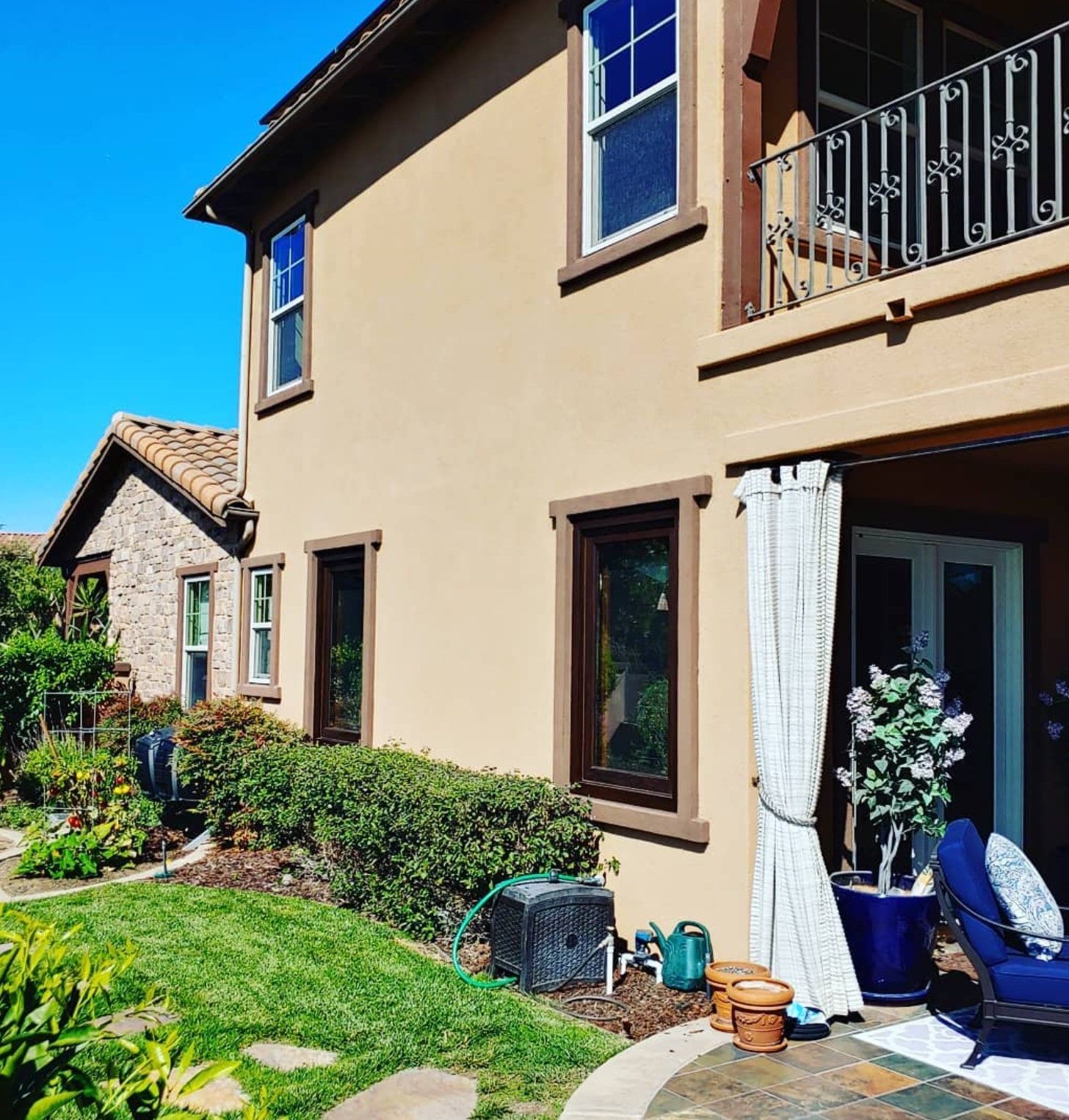 Beige two-story house with dark brown trim, green lawn, and a patio with curtains and a blue chair.