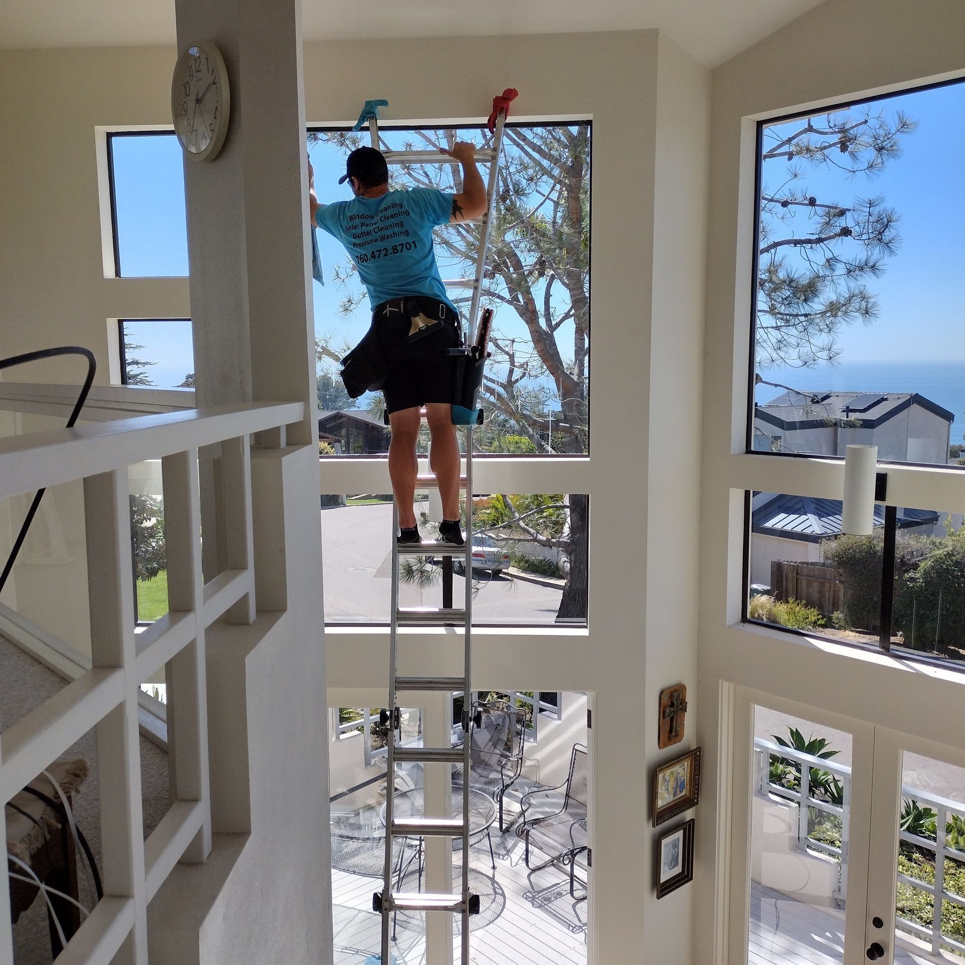 Man cleaning tall interior windows from a ladder in a two-story home with a coastal view.