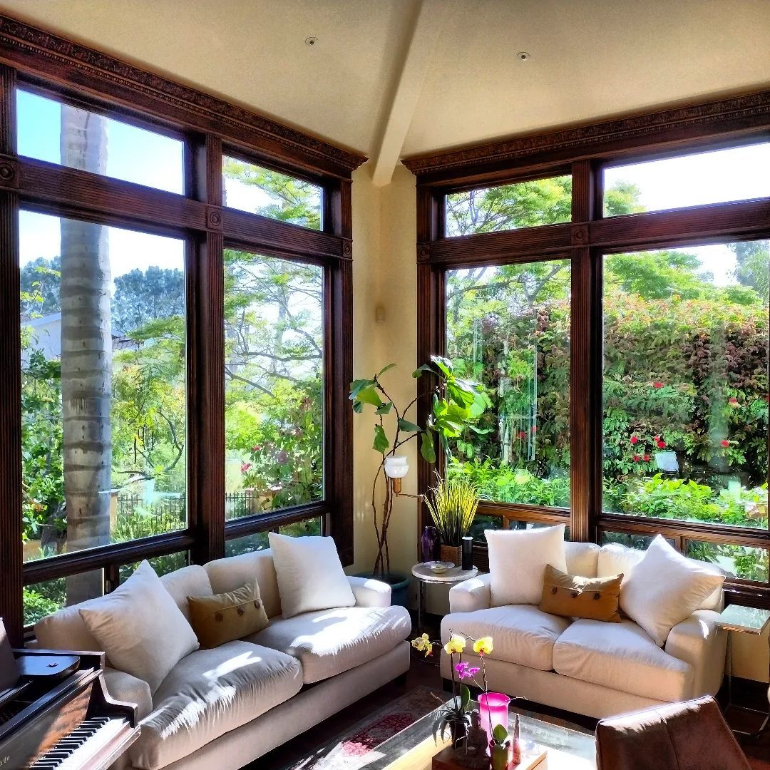 Sunlit living room with white sofas, large windows overlooking lush greenery, and dark wood trim.