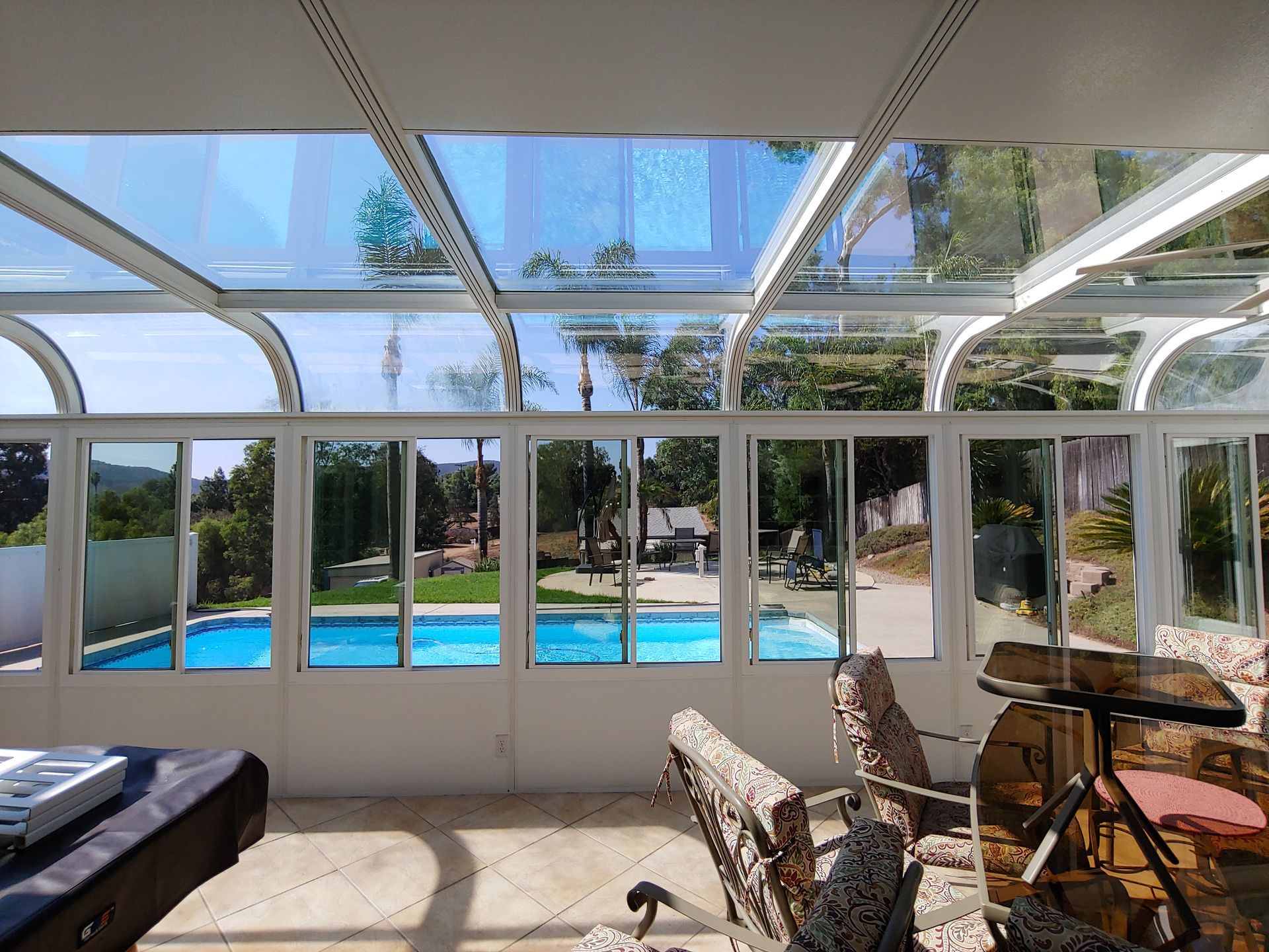 Indoor pool enclosure with glass roof and walls, overlooking a backyard pool and green trees.