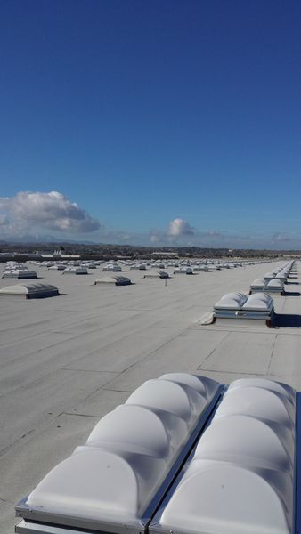 Flat commercial roof with many skylights under a bright blue sky with scattered clouds.