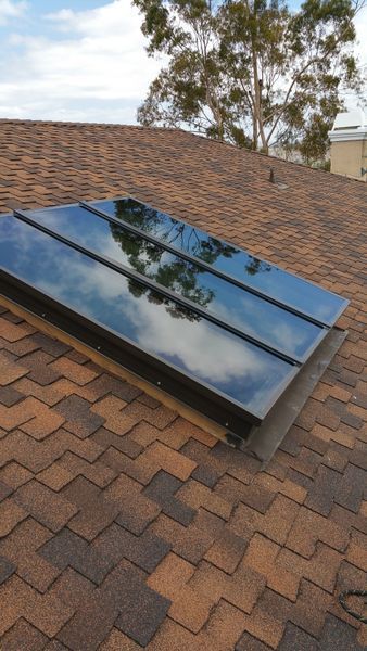 Skylight on a brown asphalt shingle roof, reflecting the sky and trees.
