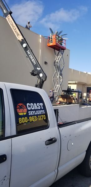 A white Coast Skylights truck with a logo. Two workers are on the roof and in a lift working on a building.