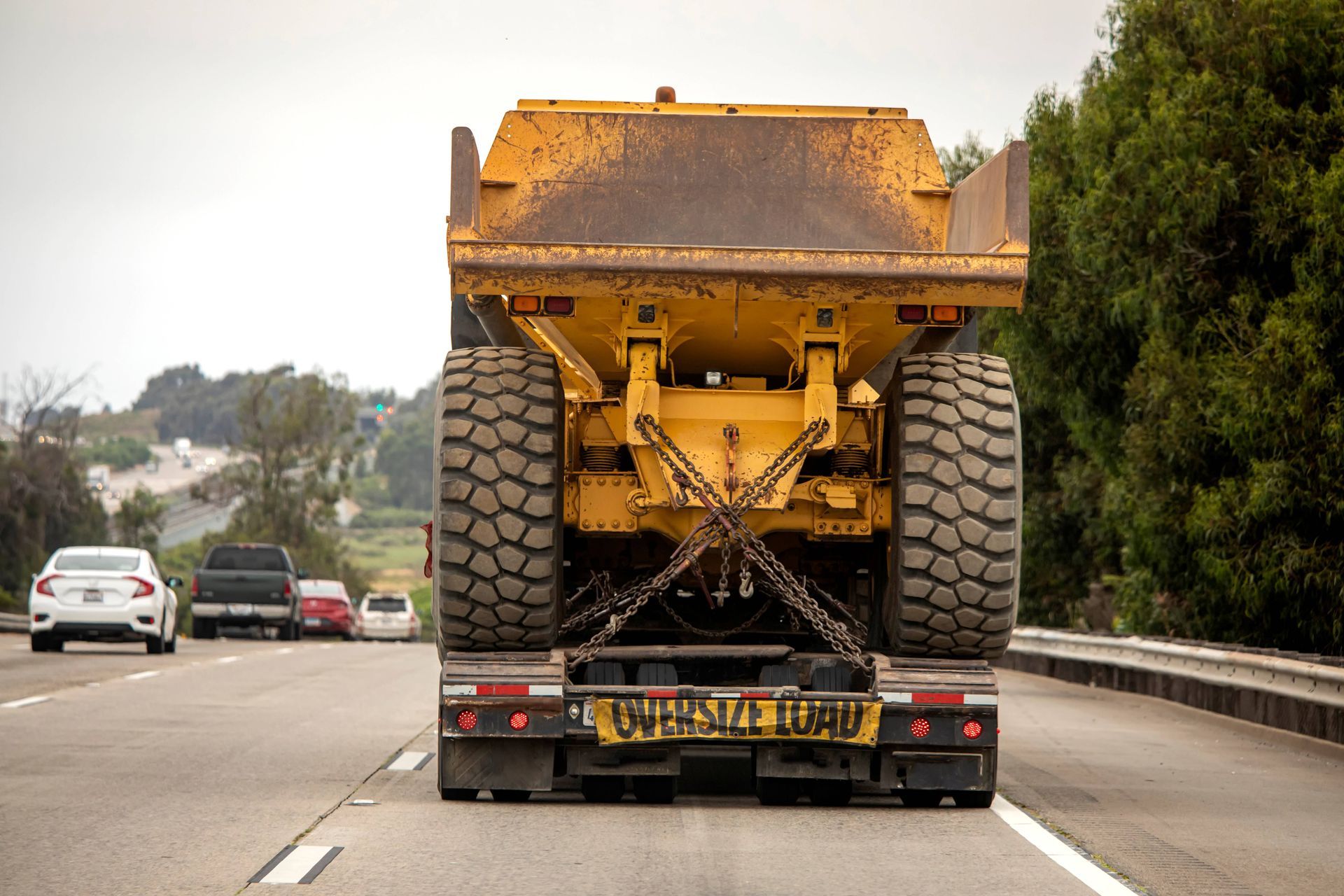 Heavy construction equipment secured on flatbed truck traveling on highway with oversize load sign.