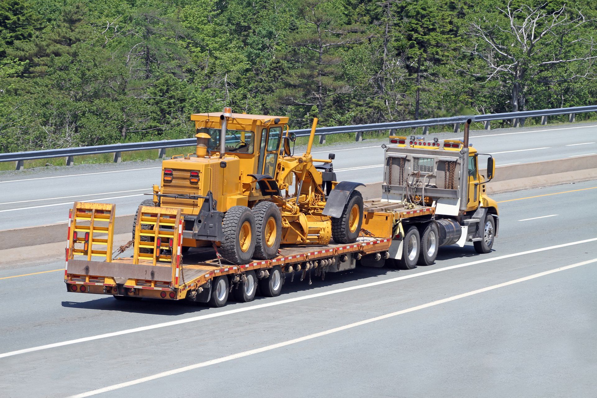 Flatbed semi truck hauling a large road grader down the highway.
