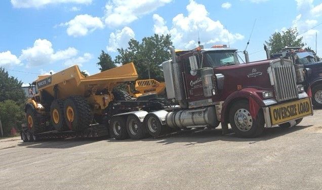 Large truck hauling an orange excavator on a flatbed trailer in a parking lot.