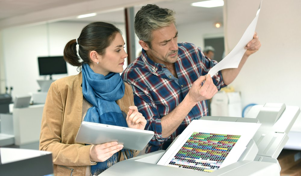Man and woman examining a printed document near a printer in an office.