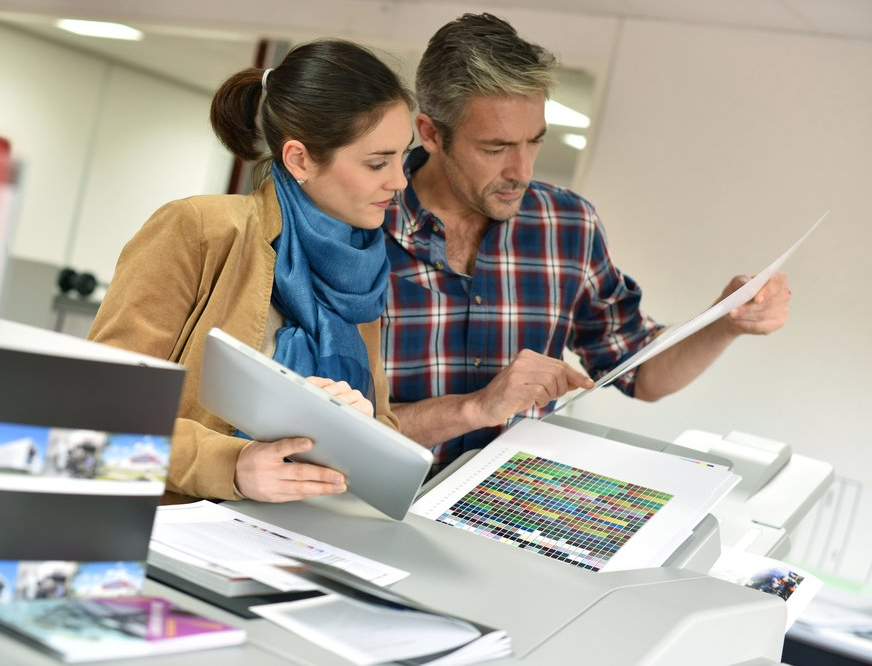 Woman and man reviewing printed documents near a printer, looking at color proofs.