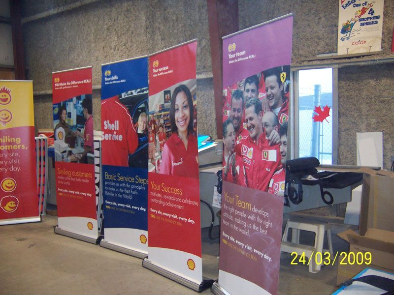 Row of Shell retractable banners in a warehouse, showing employees and promoting services in red, blue, and yellow.
