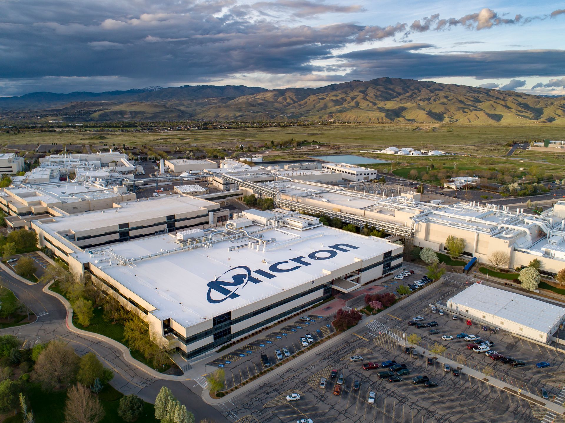 Aerial view of a large Micron factory with the company's logo on the roof and a parking lot in front.