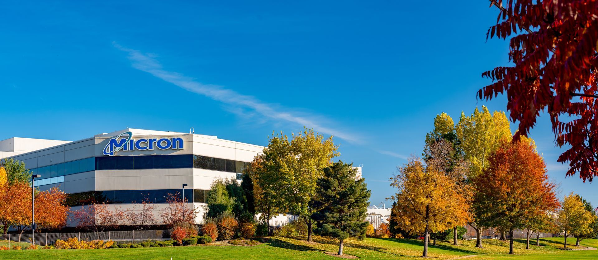 Aerial view of a large Micron factory with the company's logo on the roof and a parking lot in front.