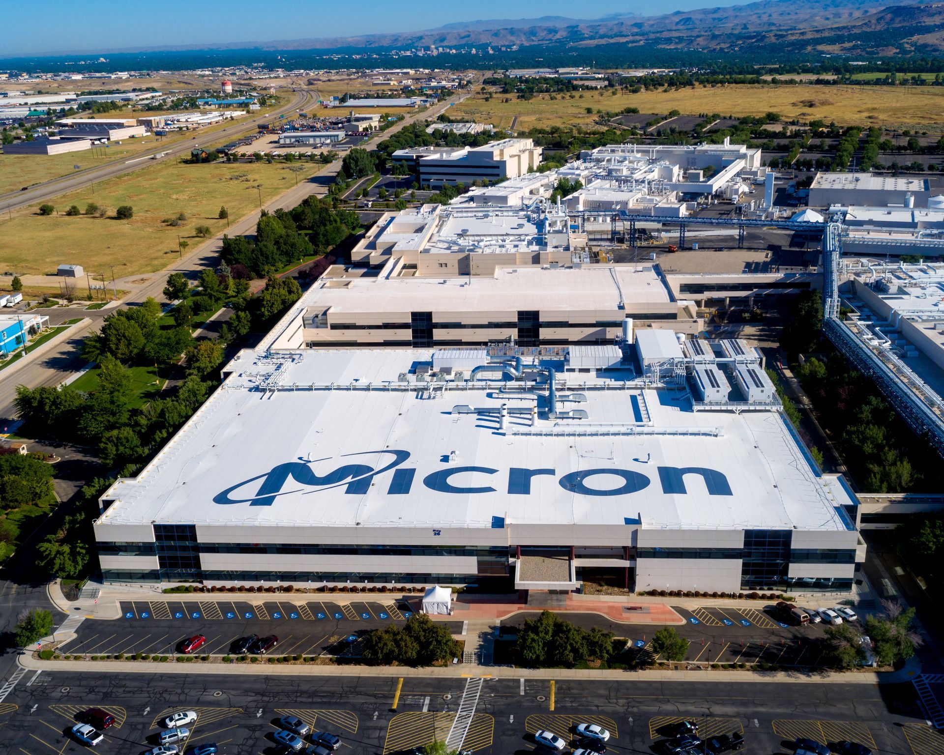 Aerial view of a large Micron factory building with the company's logo on the roof and a parking lot in front.