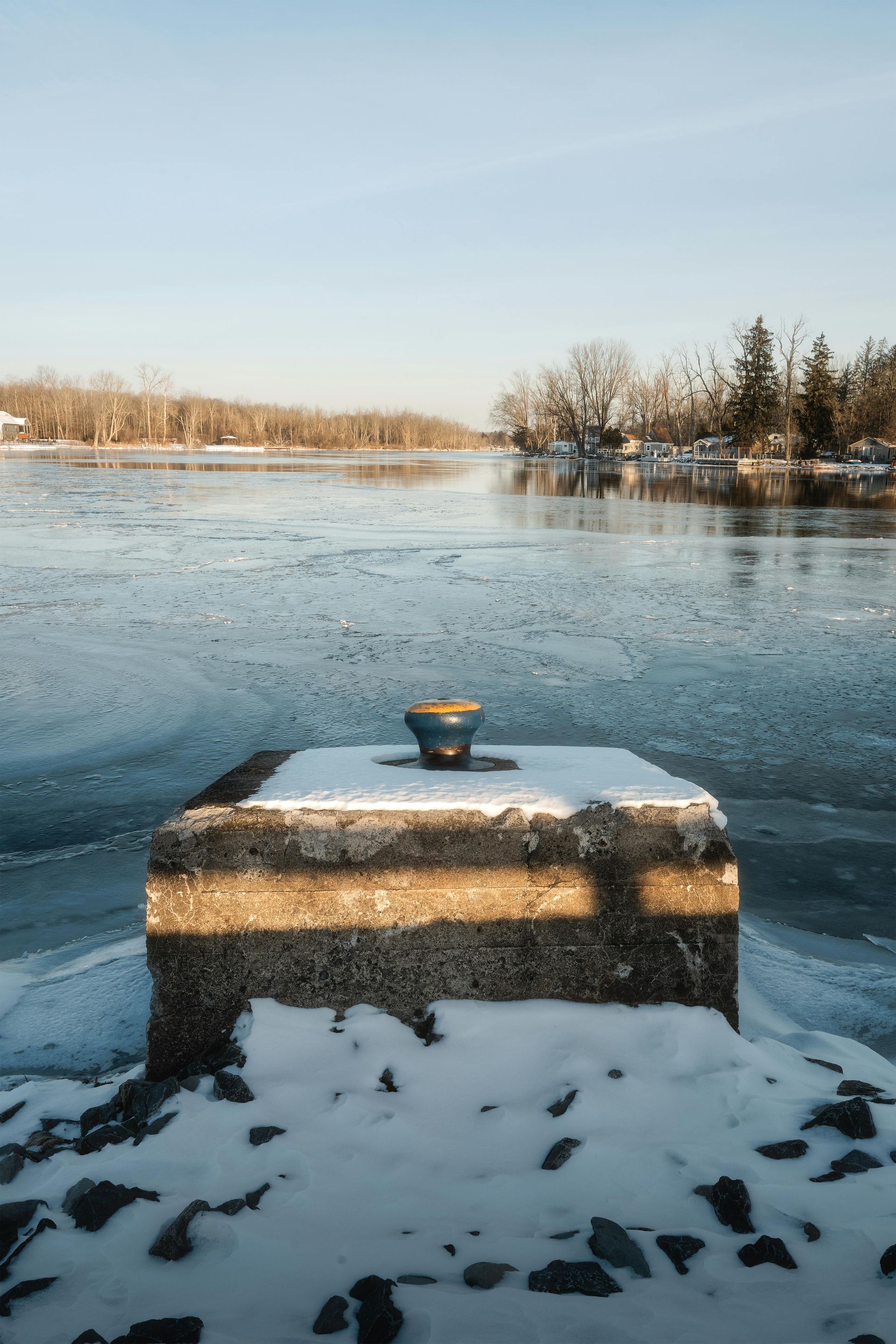 Concrete bollard covered in snow with a blue cleat sits on a frozen lake under a bright sky.