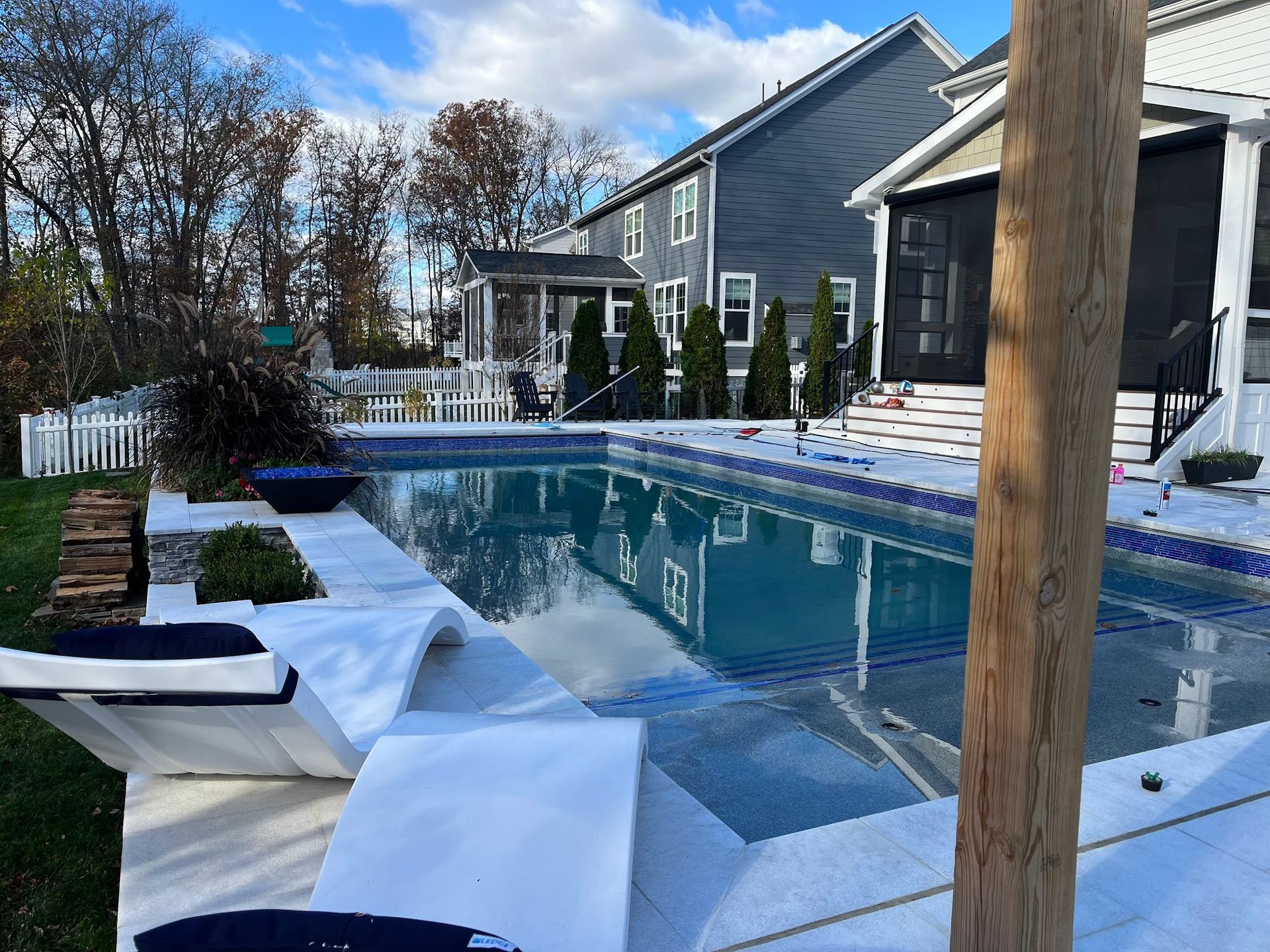 A rectangular pool with blue tile surrounded by a white patio, in front of houses and trees.