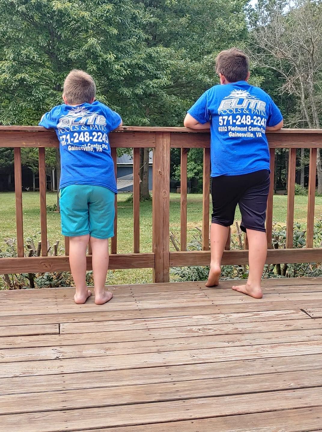 Two people in blue shirts stand on a wooden deck, looking out. Green background, sunny day.