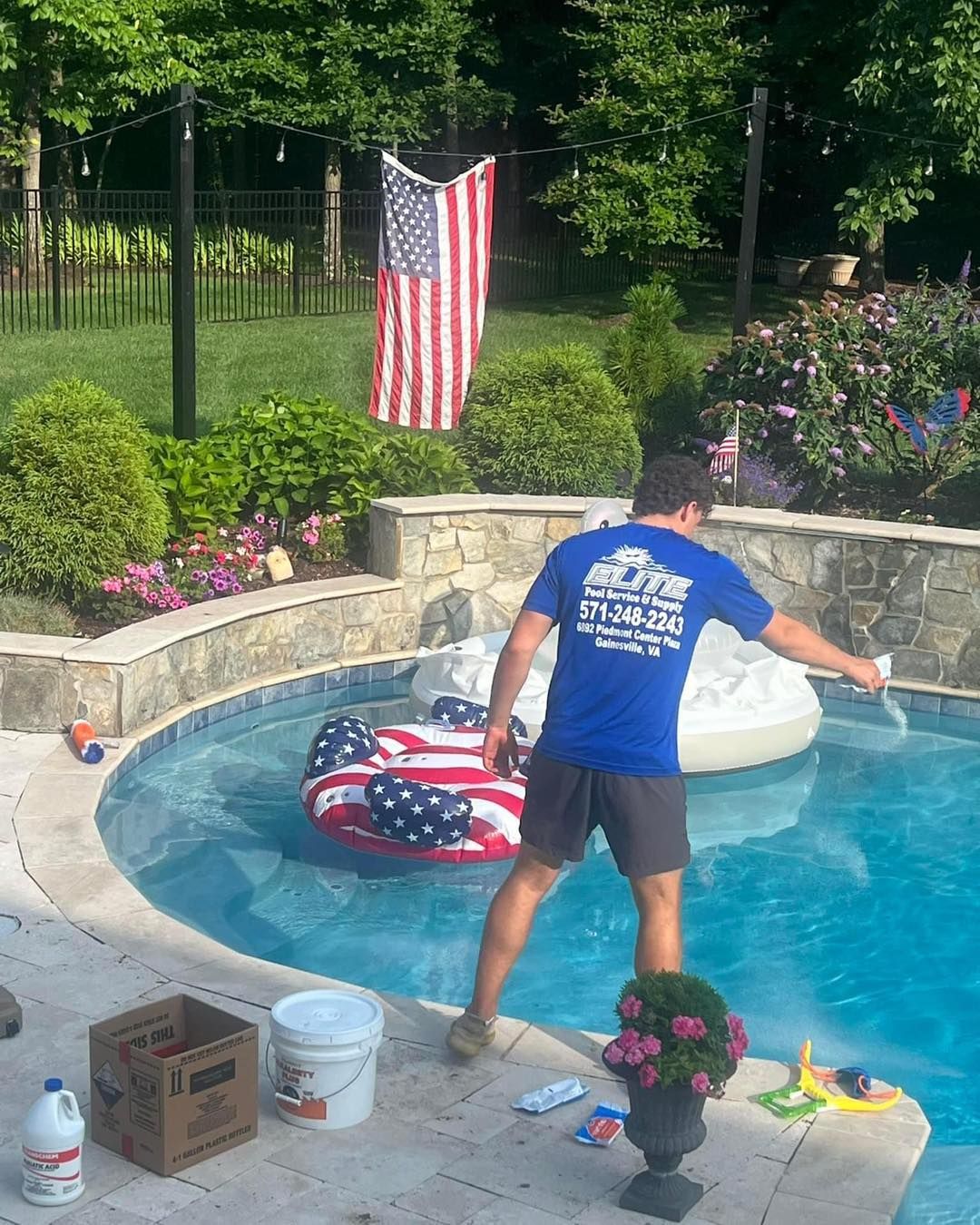 Man cleaning a pool with an American flag in the background. Pool float, chemicals, and lush landscaping are visible.